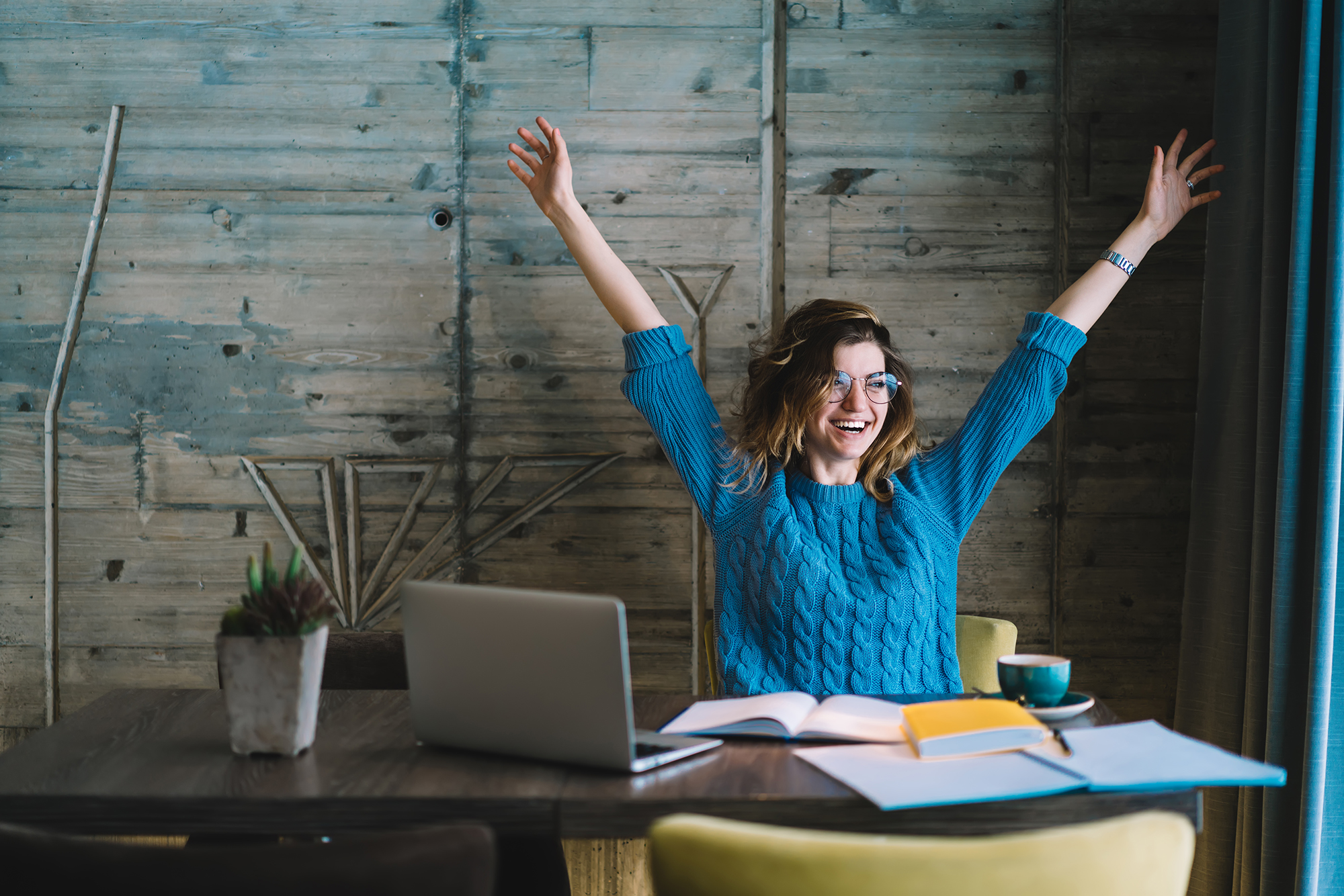 Happy hipster student rejoicing in success of training project holding hands up.Overjoyed young woman completed studying task during exam preparation sitting at desktop with laptop and textbook